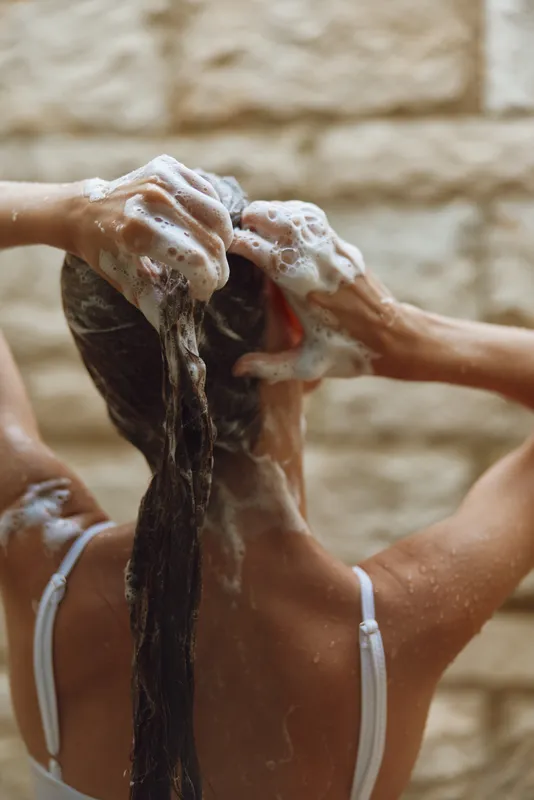 Woman washing hair with shampoo, soap foam covering wet long hair, casual white straps visible, natural warm background, personal care and hygiene concept.