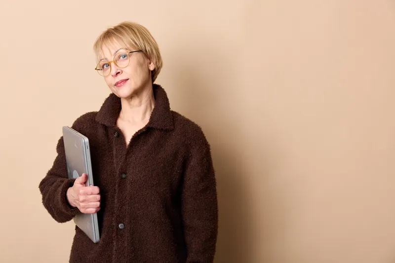 Senior woman portrait with glasses holding a notebook, calm thoughtful look, dark coat, beige studio background, reading and writing, education and planning, notes for routine