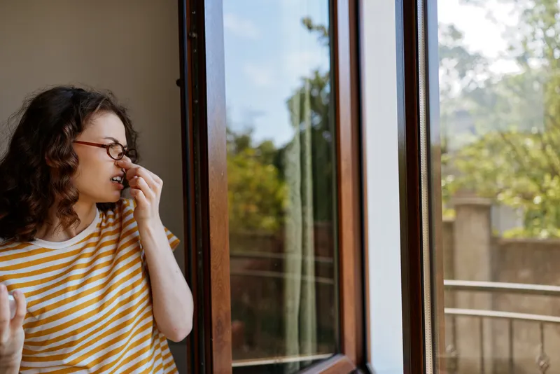 Woman with curly hair in a striped shirt stands by an open window at home, enjoying fresh air. A relaxed lifestyle scene with natural daylight and warm, casual mood.