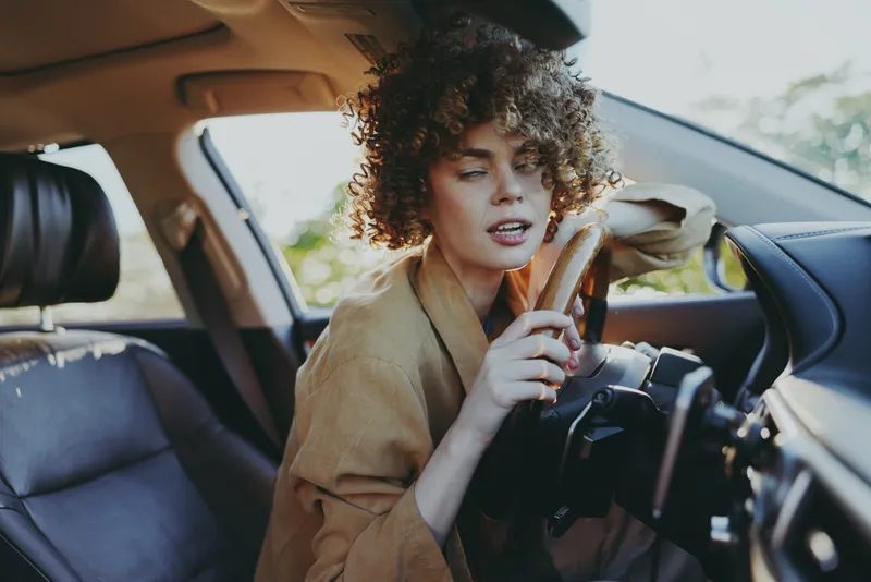 Young woman with curly hair sits in the driver's seat adjusting a camera, her focused expression framed by warm retro film color, a candid moment of creative travel.