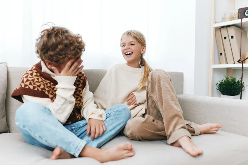 Two children sit on a light sofa, sharing a candid moment of laughter and warm connection, creating a welcoming scene of home life and carefree childhood joy.