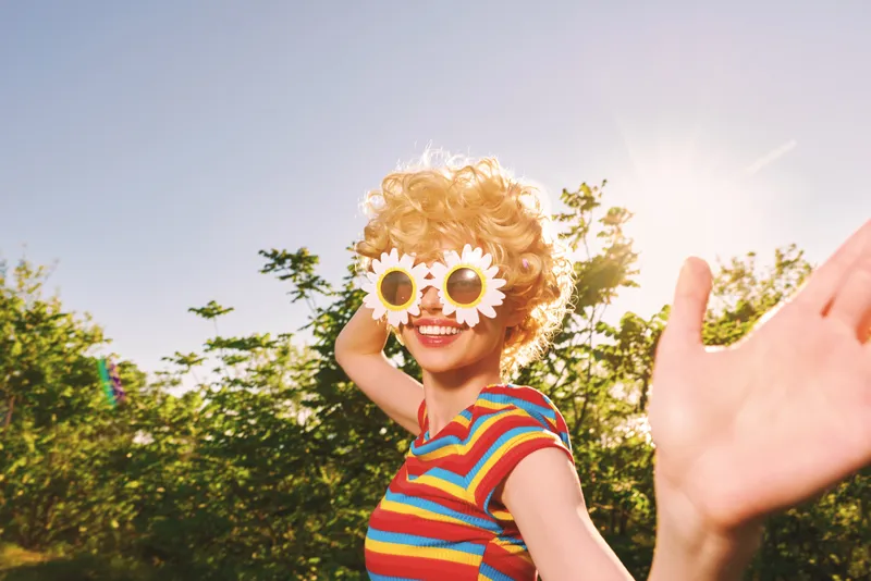 Smiling woman with flower sunglasses posing outdoors in bright sunlight, a retro film color vibe. A lifestyle moment full of summer joy, playful style, and carefree expression in a sunny garden.
