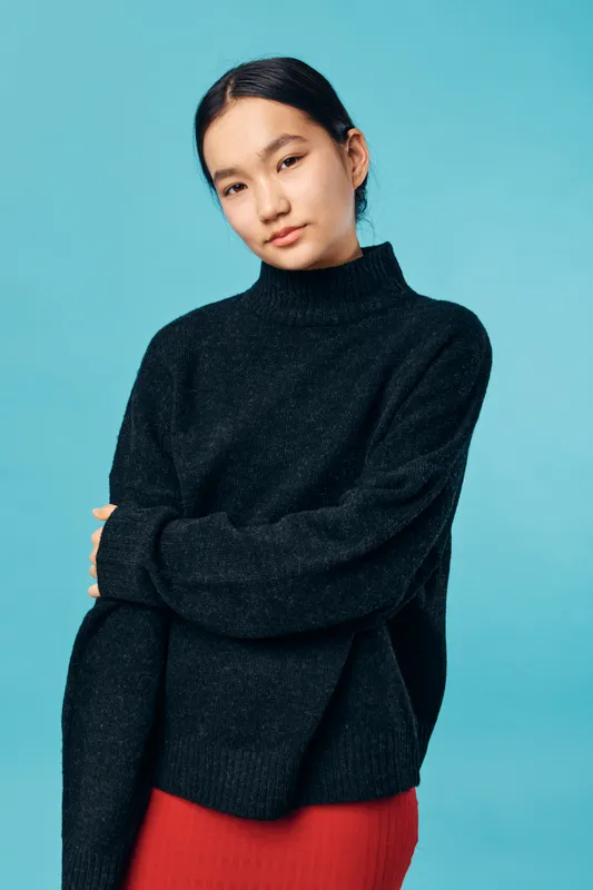 Young woman wearing black sweater and red skirt posing with arms crossed looking at camera against blue background. Casual fashion portrait with natural expression and warm lighting.
