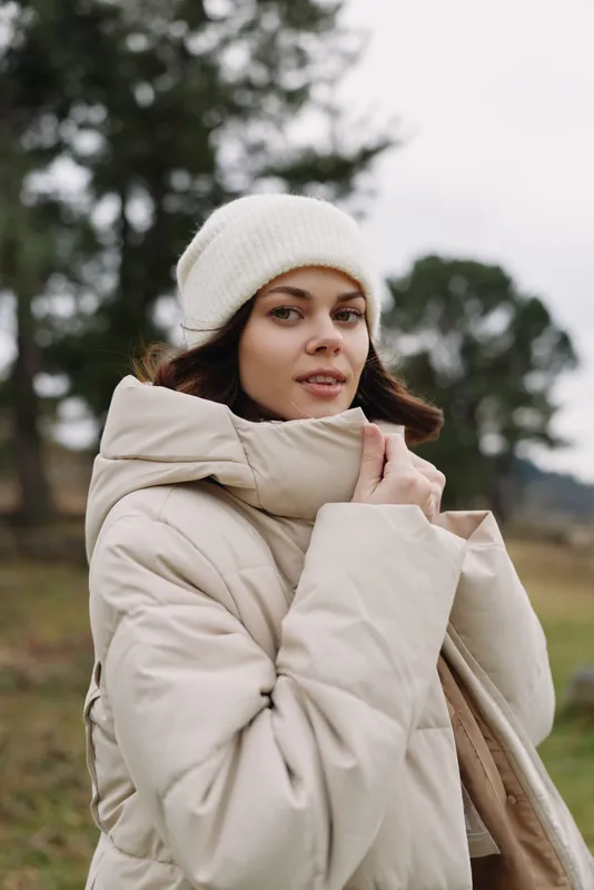 Woman wearing a thick winter coat and beanie stands outdoors in a quiet countryside, ready for cold weather exploration and fresh air under soft, overcast skies
