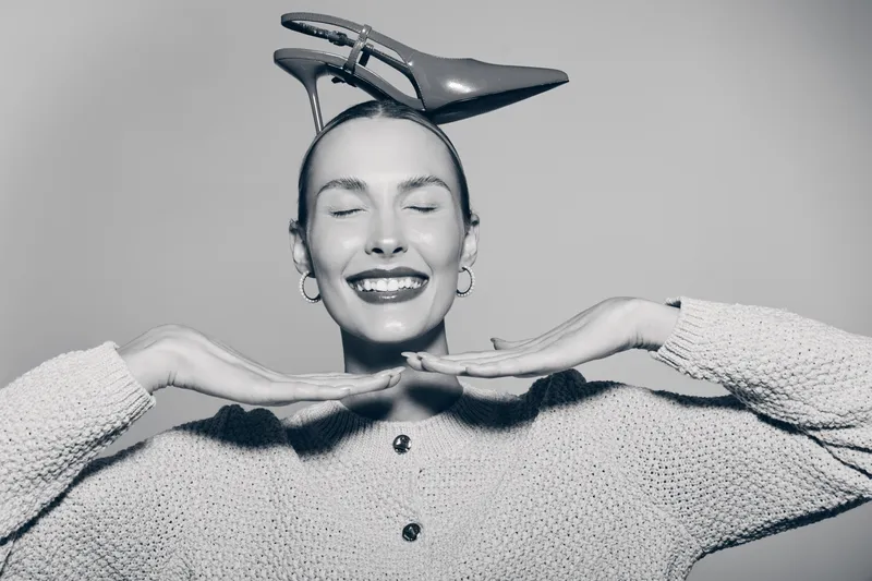 Happy white woman with light skin smiling with closed eyes and high heel shoe on head, wearing cozy sweater against plain background.