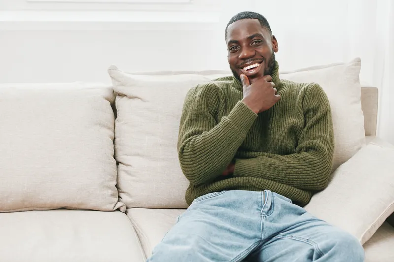 Smiling young man in a cozy living room wearing a green sweater and jeans, sitting comfortably on a beige couch, embodying tranquility and joy
