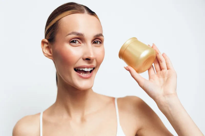Smiling young woman with clear skin holding golden cosmetic jar close to face isolated on white background showing skincare product and positive facial expression in beauty concept.