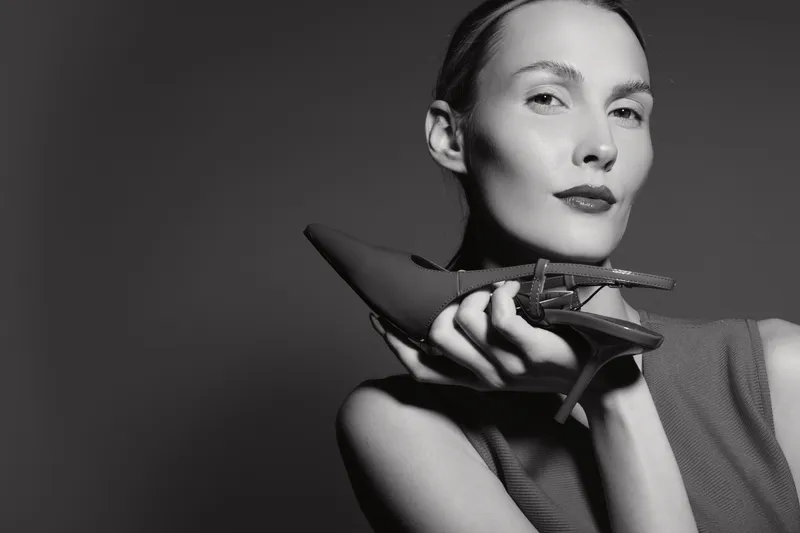 Stylish white woman with light skin holds high heel shoe near face in black and white studio portrait. Concept of fashion, elegance, and modern style.