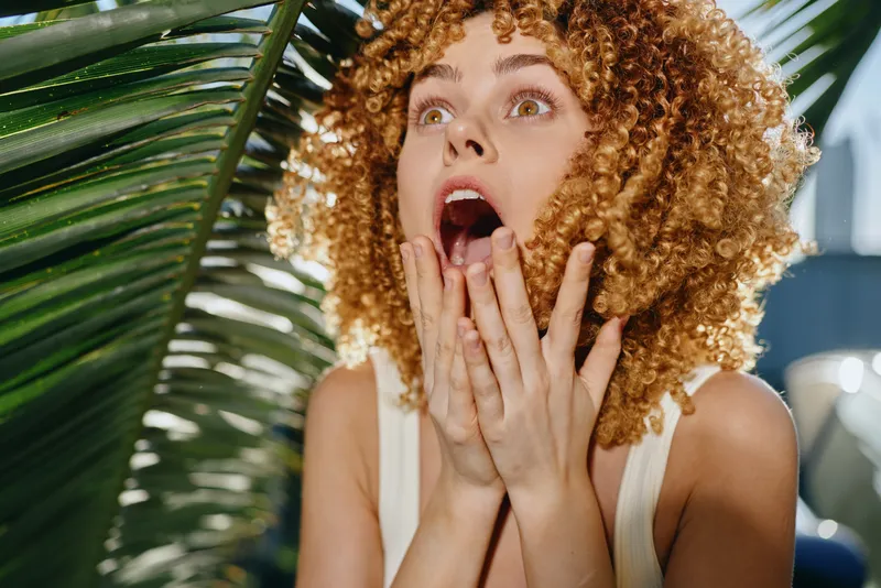 surprised woman with curly hair expressing shock and amazement outdoors near tropical green leaves in bright sunlight, natural emotion in casual summer outfit.