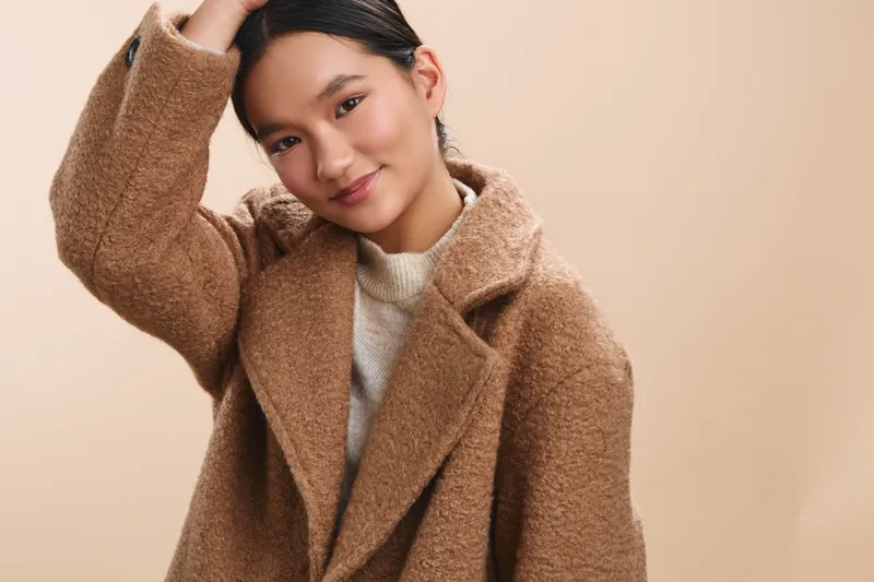 Asian woman wears a warm brown coat in a neutral studio setting, her relaxed pose and gentle smile conveying approachable style and cozy confidence.