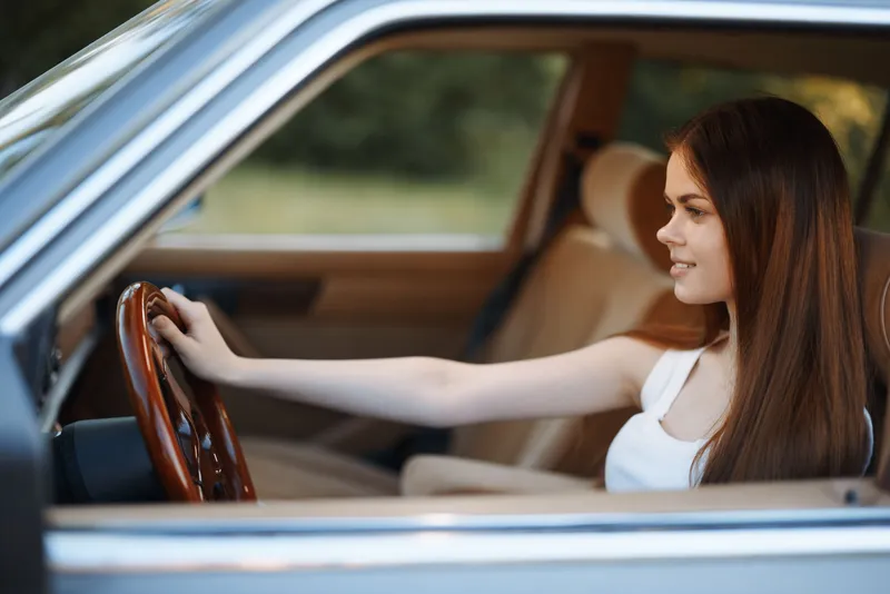 Young woman driving a vintage car, smiling cheerfully with long brown hair, dressed casually in a white tank top, in a sunlit outdoor setting.