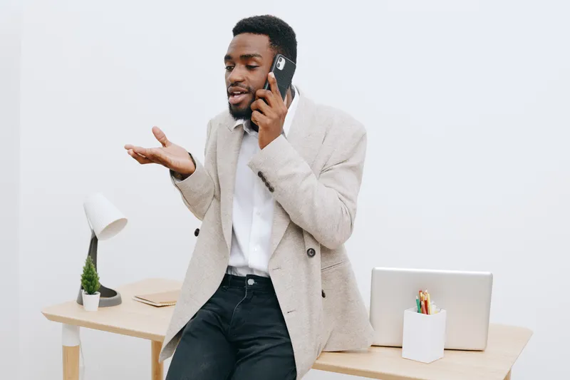 Confident young African American man wearing smart casual outfit talking on mobile phone in modern office space, gesturing with hand, smiling, isolated on plain white background, professional lifestyl