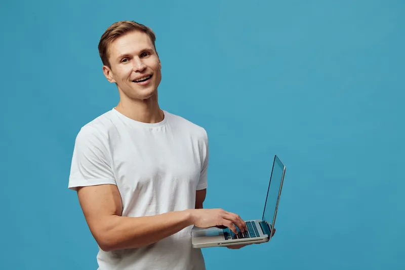 Young smiling man holding a laptop in hand, wearing a white t-shirt, isolated on a solid light blue background, studio portrait, casual attire, technology, communication, cheerful mood, people lifesty