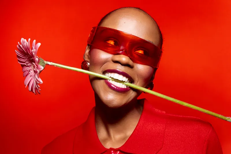 Smiling black woman with dark skin wearing red sunglasses and red shirt holds pink flower in mouth. Bright red background creates vibrant, playful mood. Concept of joy, fashion, and beauty.