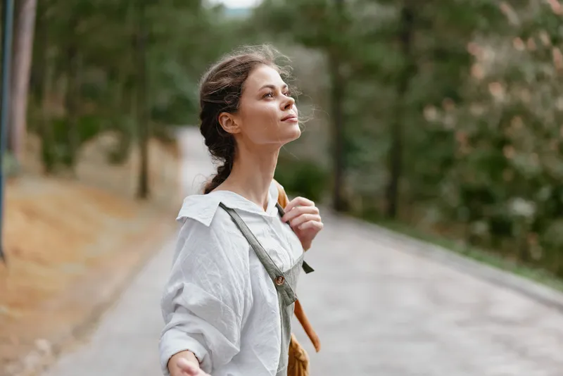 Young woman enjoying a serene moment outdoors, dressed in a casual white shirt with earthy tones, expressing joy in a tranquil forest setting