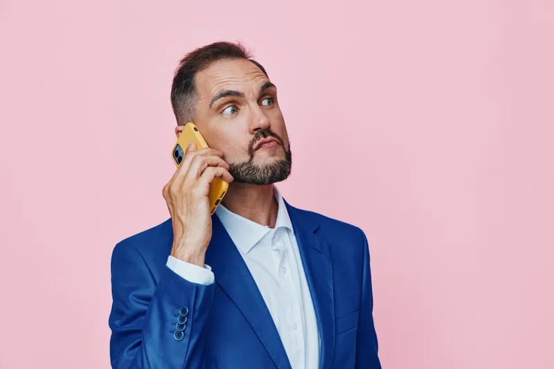 A professional man in a blue suit speaks on a phone against an isolated colored pink background, exuding confident business demeanor and calm, focused emotion.