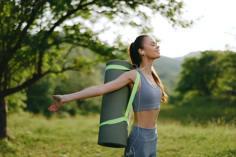 A joyful young woman enjoying outdoor yoga, carrying a mat in a serene green landscape, showcasing fitness, wellness, and nature, ideal for healthy lifestyle concepts