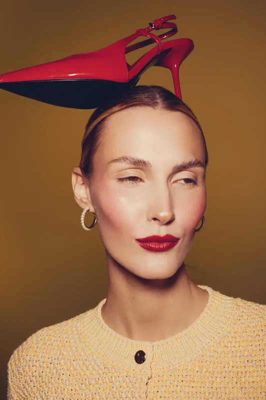 Portrait of a woman with red lipstick and hoop earrings balancing a red high heel shoe on her head against a brown background. Concept of fashion, style, and creativity.