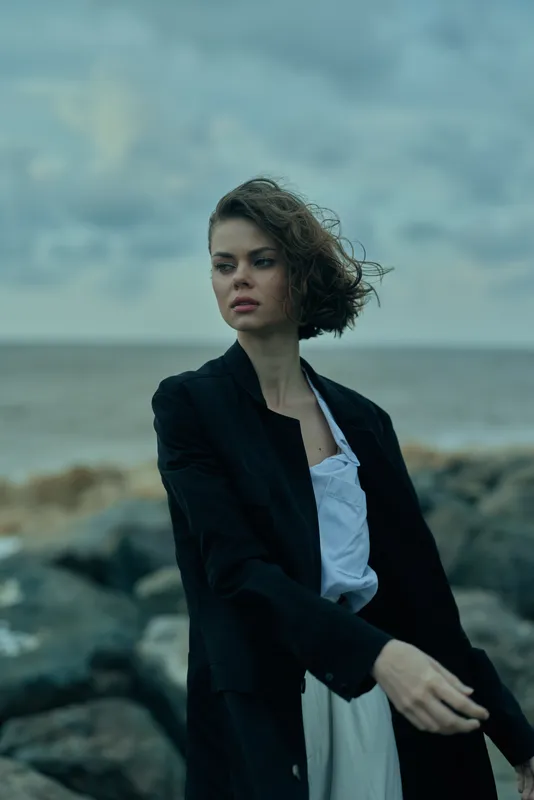 Woman in a black blazer standing on a rocky coast by the sea, windswept hair and thoughtful expression, minimalist fashion shoot with moody ocean backdrop, natural light highlighting tailored