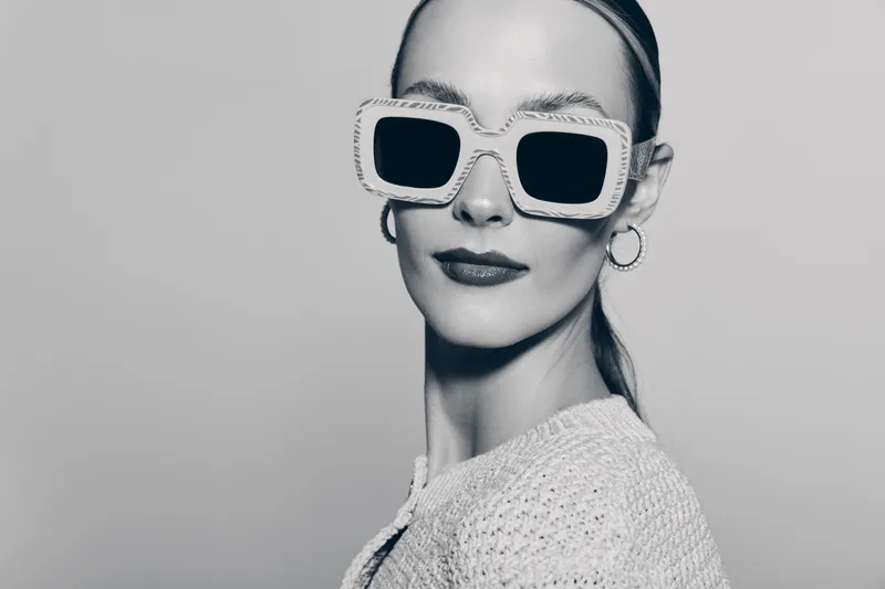Stylish white woman with pale skin wearing large square sunglasses and hoop earrings poses confidently in knitted top against neutral background. Fashion and elegance concept.