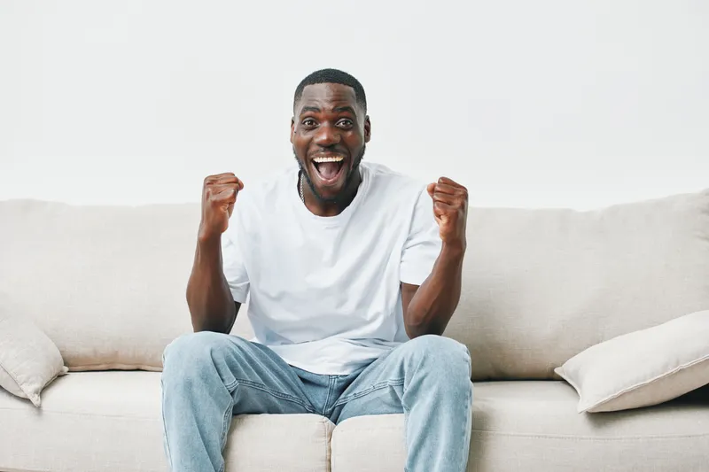 Joyful young African man celebrating victory on a comfortable sofa, wearing casual white t shirt and jeans, expressive emotions in bright and minimalistic interior