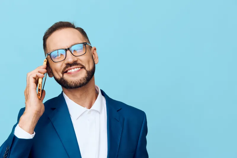A business professional man on the phone, confident and pleased, standing against a solid blue background, projecting focus, success, and clear communication.