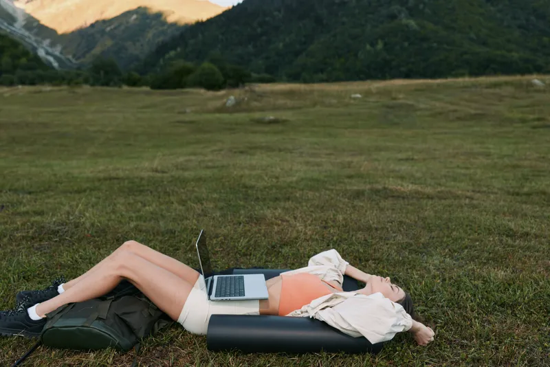 A relaxed professional lies on a grassy field with a laptop resting on the torso, sunlit mountains in the distance, conveying remote work and leisure outdoors.