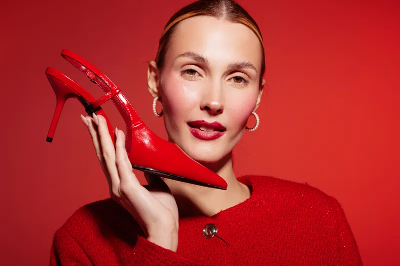 Woman in red sweater holds red high heel shoe near face against red background. Fashion concept with matching colors, style, and confident female model posing in studio lighting.