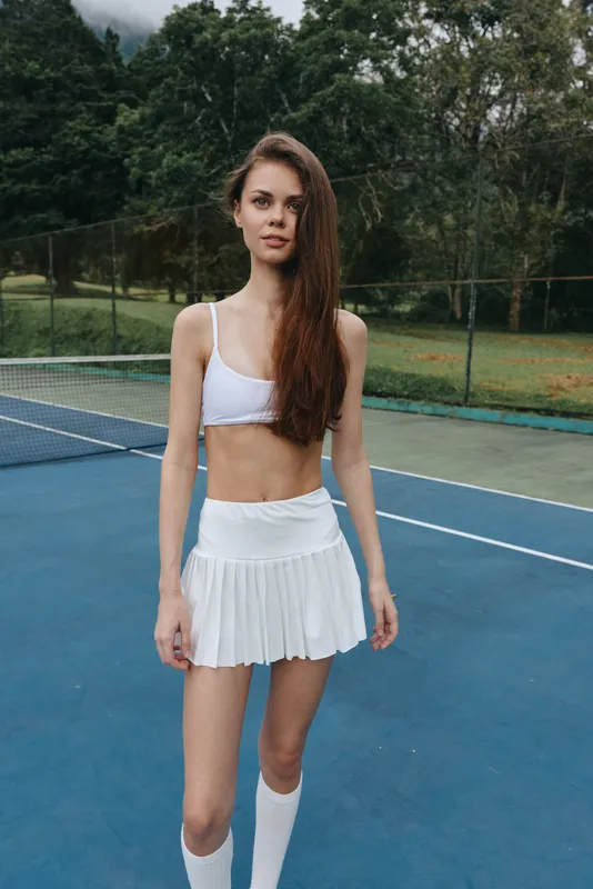 Young female tennis player on the court in a white skirt and sports bra, exuding confidence and athleticism in a natural green background