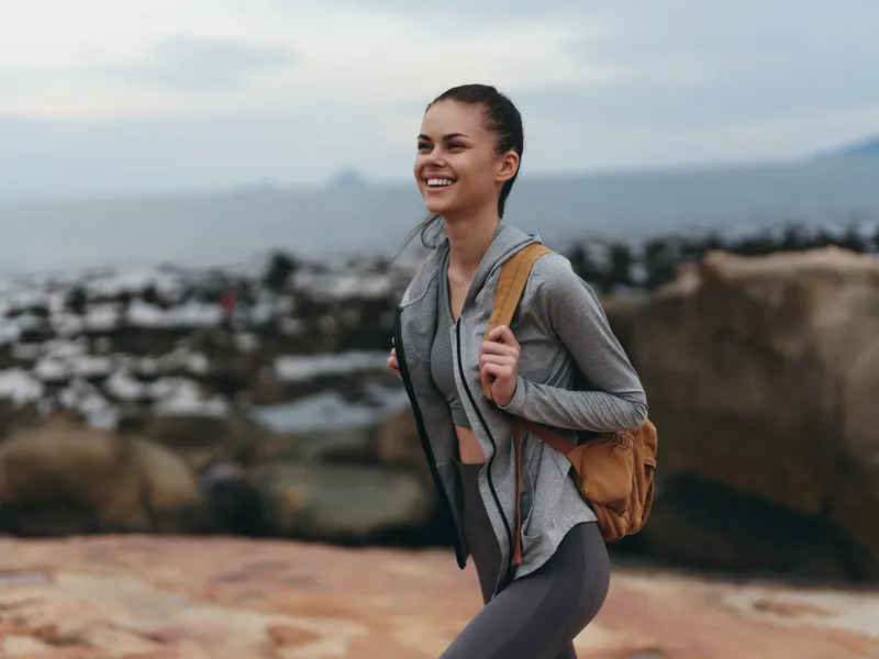 Cheerful female hiker in athletic wear enjoying nature with a backpack, rocky seaside background, showcasing a healthy lifestyle and adventure spirit