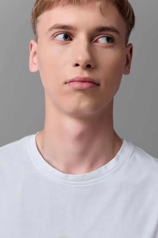Young man in a white T shirt with a neutral expression, gazing to the side, in a light gray studio setting, minimal lifestyle image without branding and distractions.