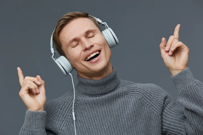 Joyful young man enjoying music wearing headphones, closed eyes, smiling, casual gray turtleneck sweater, isolated on plain dark background, lifestyle concept, capturing happiness, relaxation, and mus
