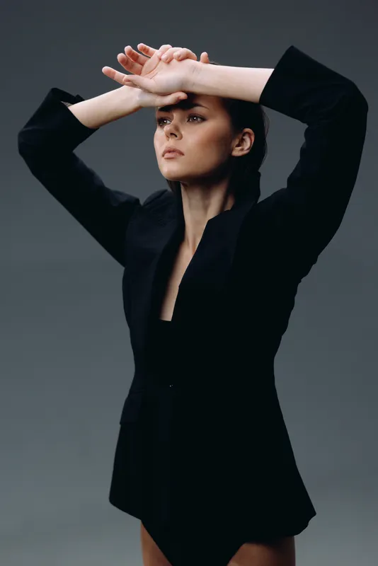 Serious woman in black jacket posing with raised arms against gray background, showing confidence and elegance.