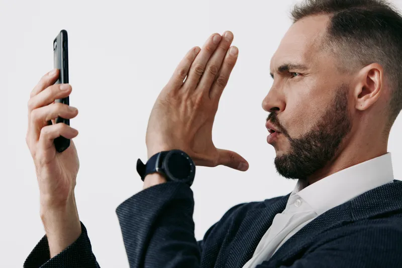 A focused business professional stands against a clean isolated colored background, showing determination and curiosity as he interacts with a mobile device.