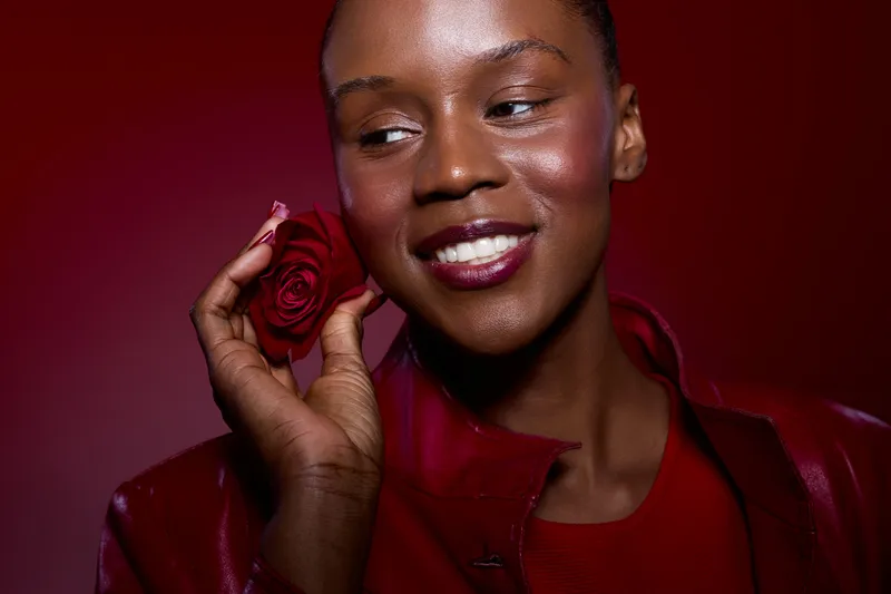 Smiling African woman with dark skin holding red rose near face, wearing red leather jacket and dress. Studio portrait with red background. Concept of beauty, elegance, and natural skincare.