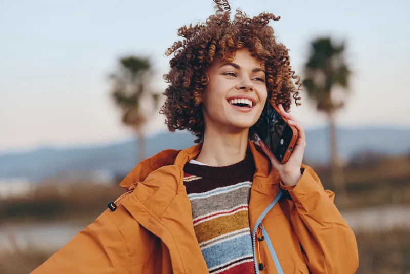 Woman lifestyle portrait outdoors, smiling with curly hair and orange jacket, one hand near headphones, sunset glow over lake and palm trees, relaxed travel mood, natural light today.