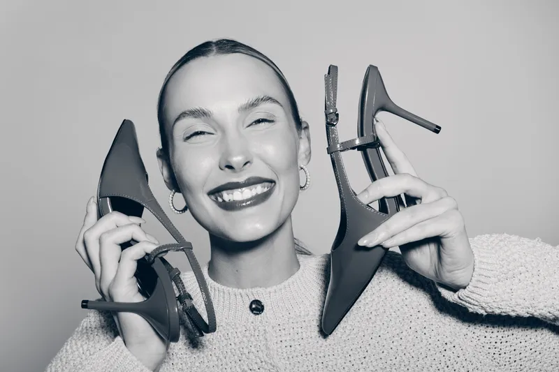 Young white woman with fair skin smiling broadly holding two high heel shoes near her face. Concept of fashion, happiness, and style.