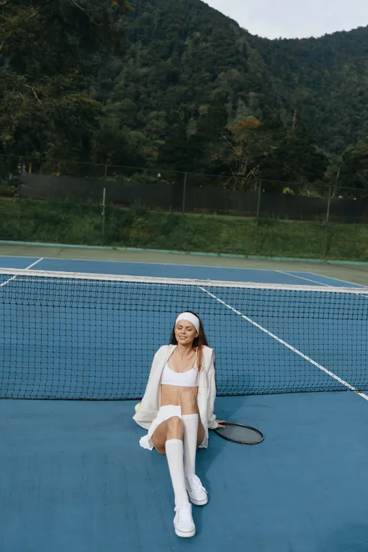 Young female tennis player in white sportswear, relaxed on the court, conveying a sense of calmness and athleticism against a green nature backdrop