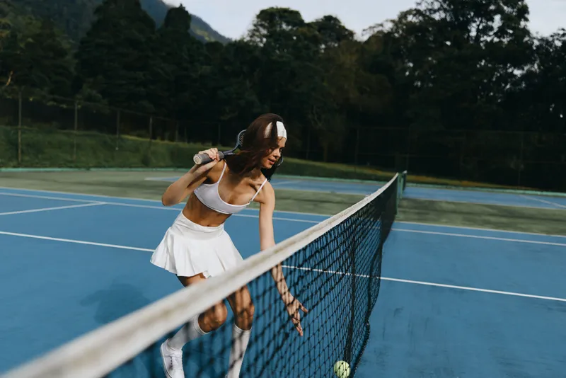 Young female athlete in white sportswear, playing tennis on an outdoor court, expressing concentration and dedication, surrounded by a vibrant green landscape