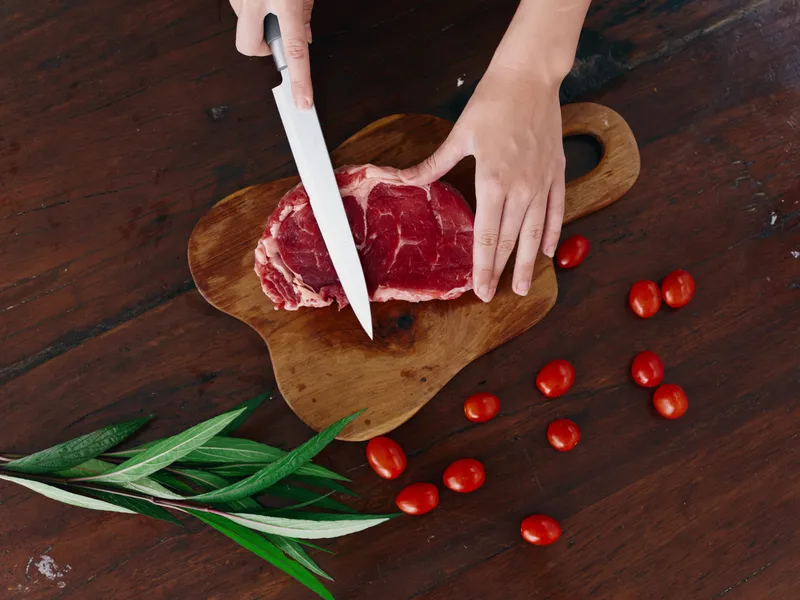 Женщина с ножом в руке нарезает свежий стейк из мяса для жарки на кухне, рядом соль, перец и другие специи на столе, красные помидоры черри и зелень, приготовление ужина