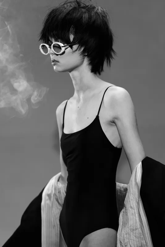 Black and white portrait of a stylish woman wearing sunglasses and a swimsuit with a relaxed expression and layered clothing