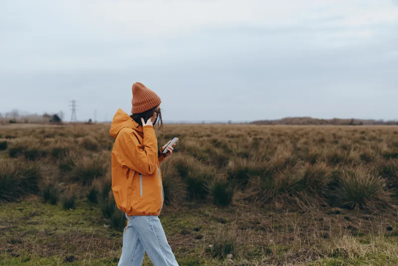 Woman lifestyle outdoors, walking in grassy field on autumn day, orange jacket and knit hat, holding phone, side view, calm mood, cloudy sky, natural light