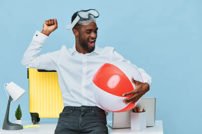 Happy young black man in white shirt enjoying fun with inflatable beach ball at office desk with suitcase and desk lamp on light blue background, cheerful mood, leisure concept