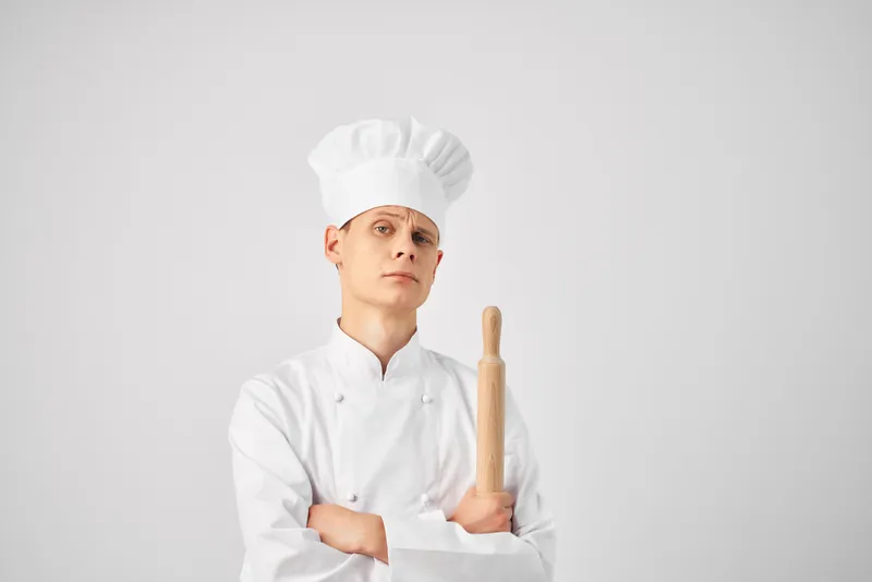 Мужчина в поварской форме держит скалку, готовит еду на кухне ресторана