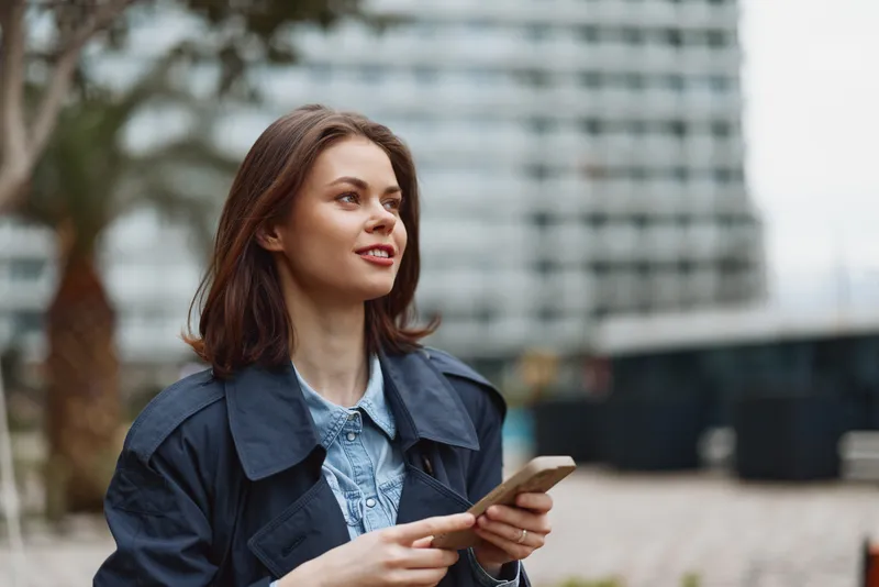 Young woman in a denim jacket holds a smartphone outdoors, glancing sideways with a soft smile, urban background and natural light creating a relaxed, candid moment.