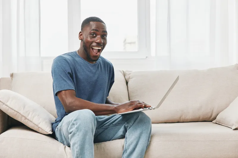 Happy young African man using laptop at home, expressing joy while sitting on beige sofa, bright natural light in a cozy living room