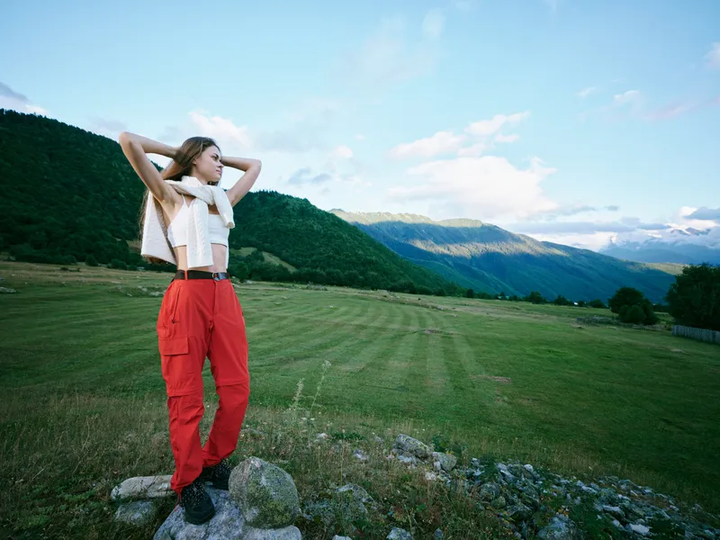 Outdoor fashion model in a mountain field wearing red pants and white top, hands raised, carefree pose under blue sky, showcasing nature inspired style and rural landscape