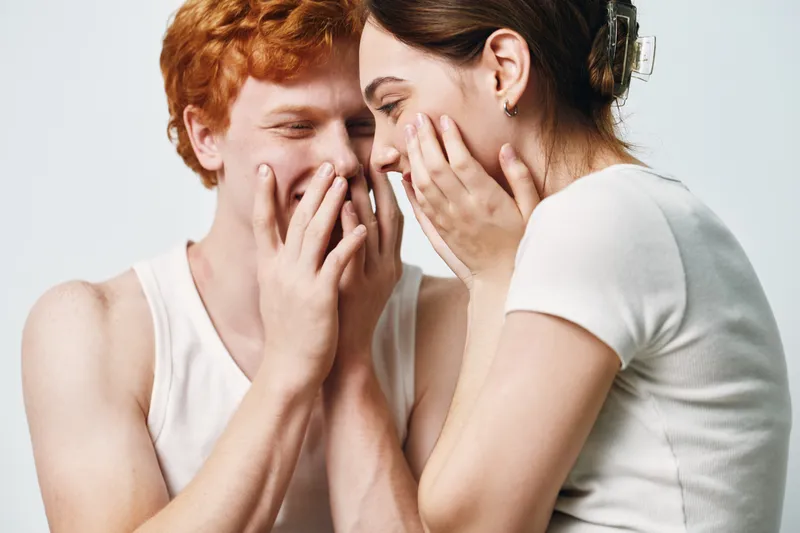 Young man and woman whispering and smiling with hands near mouth, closeup portrait on white background. Playful communication and secret sharing between friends indoors.
