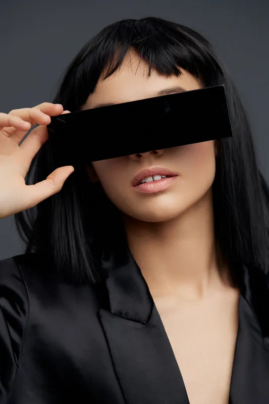 Женщина в черном костюме держит черные солнцезащитные очки перед глазами и смотрит в камеру с серьезным выражением лица