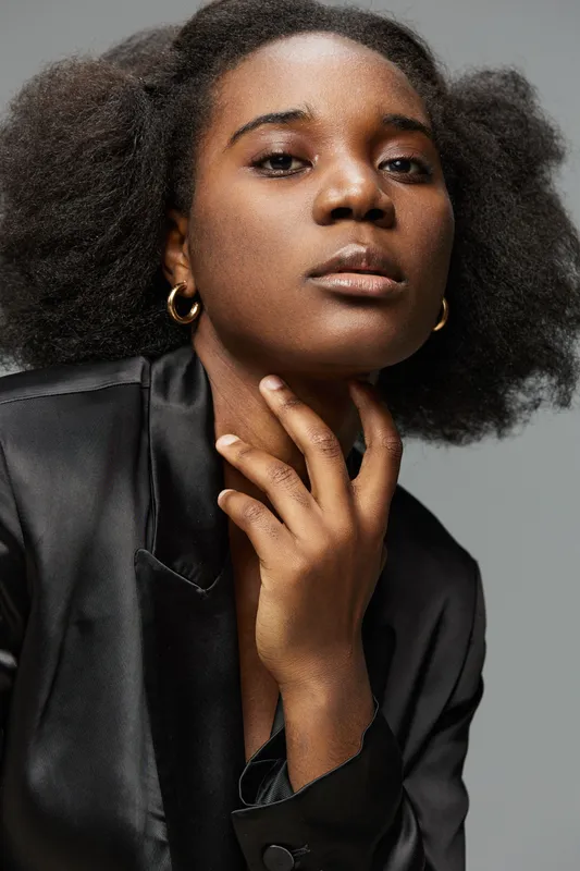 Elegant black woman in a stylish satin blazer, showcasing a confident expression with natural curly hair against a soft gray background